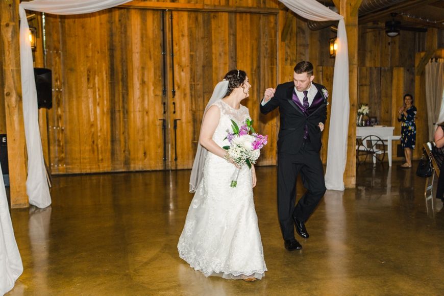 A newlywed couple walks through a rustic barn venue decorated with white draping.