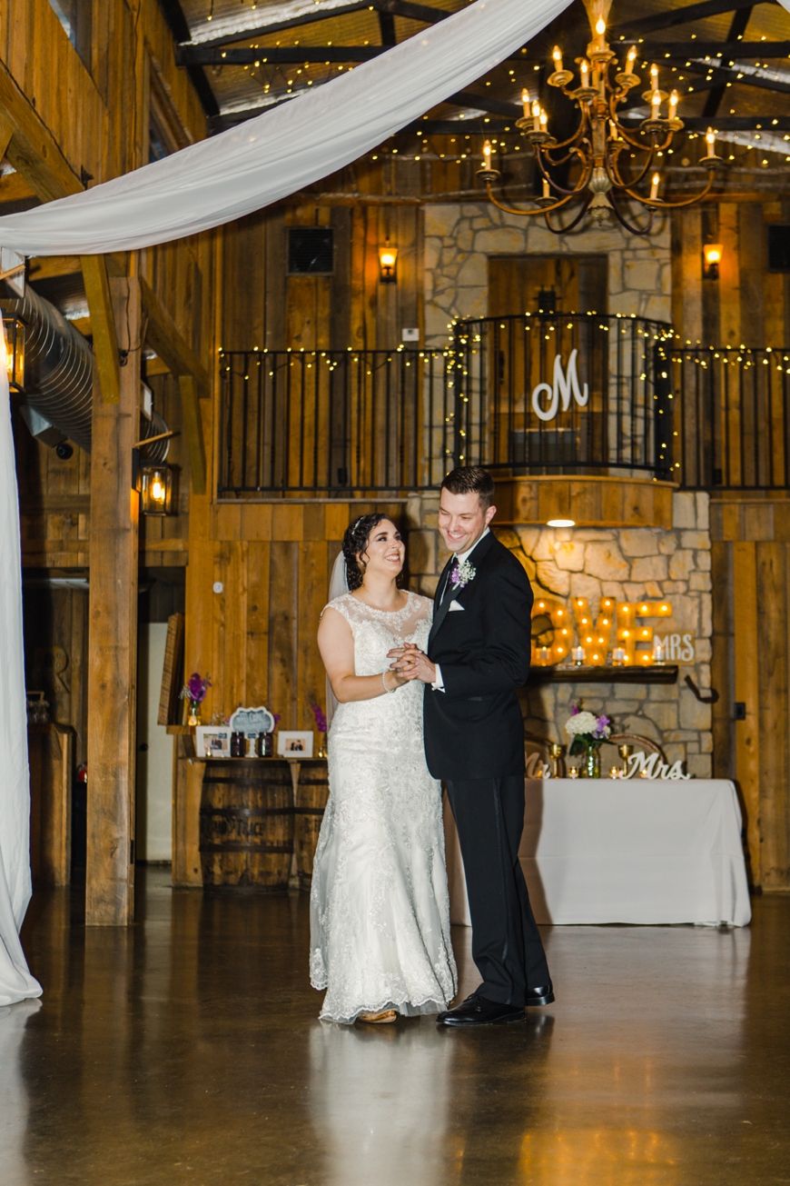 A couple dances in an elegant rustic venue with stone fireplace and chandelier lighting.