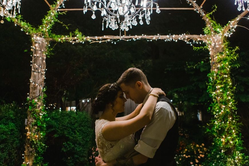 Romantic nighttime wedding photo under twinkle lights and greenery archway.