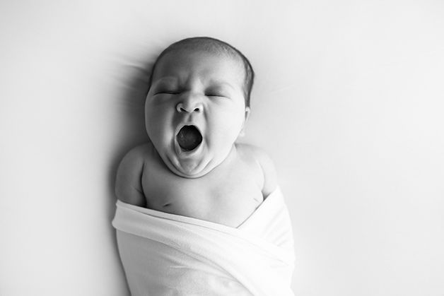 Black and white image of newborn baby girl on white blanket, yawning.