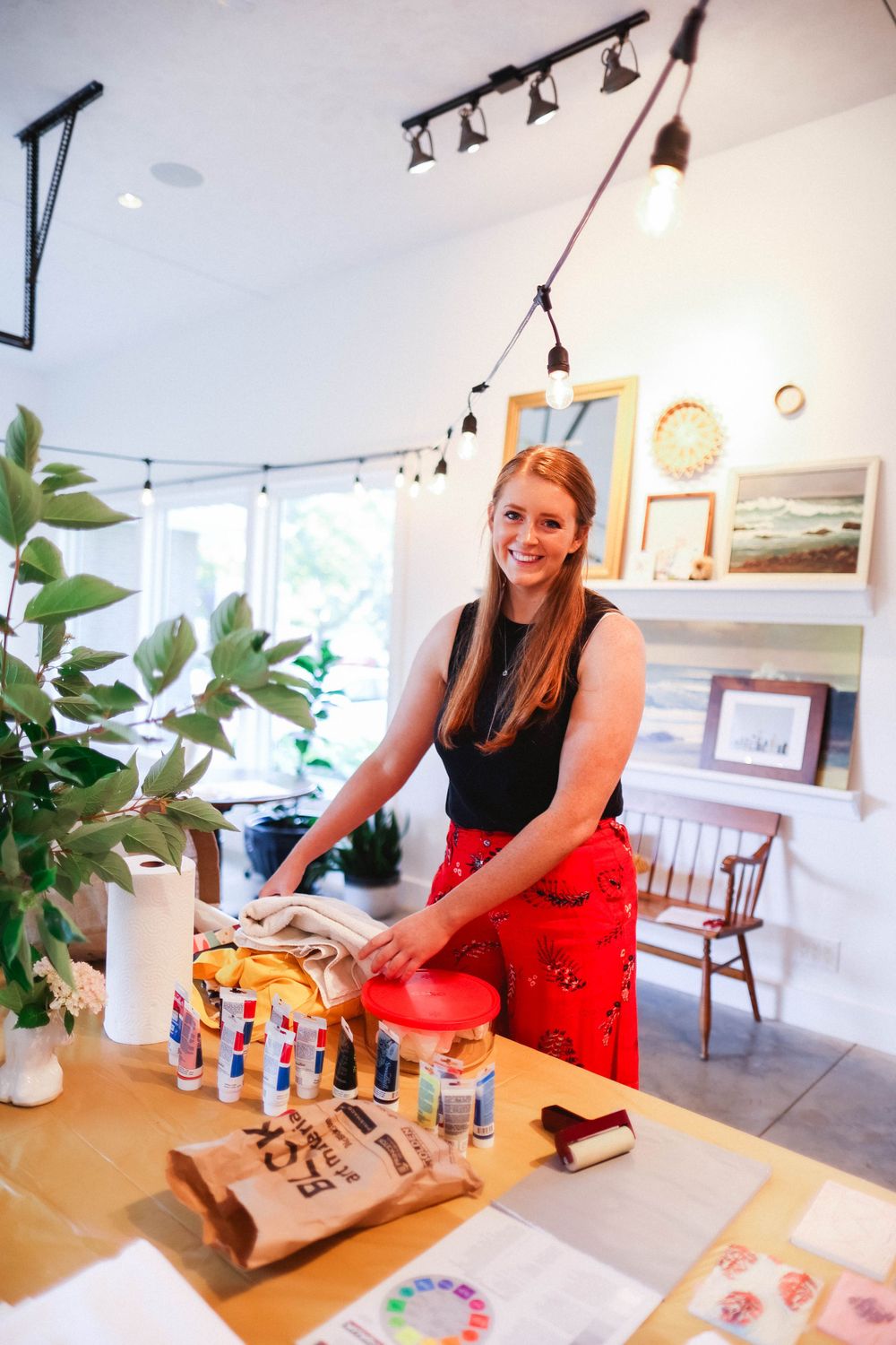 Person in red dress working with art materials at a wooden table in a gallery setting.