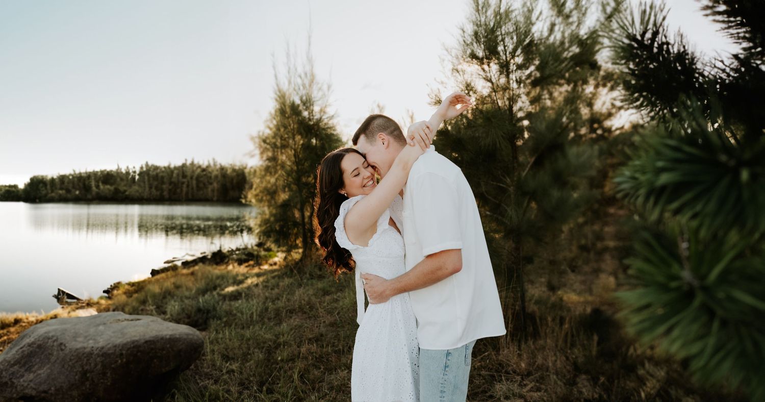 Couple embraces in romantic sunset photoshoot by a serene lakeside with trees in background.