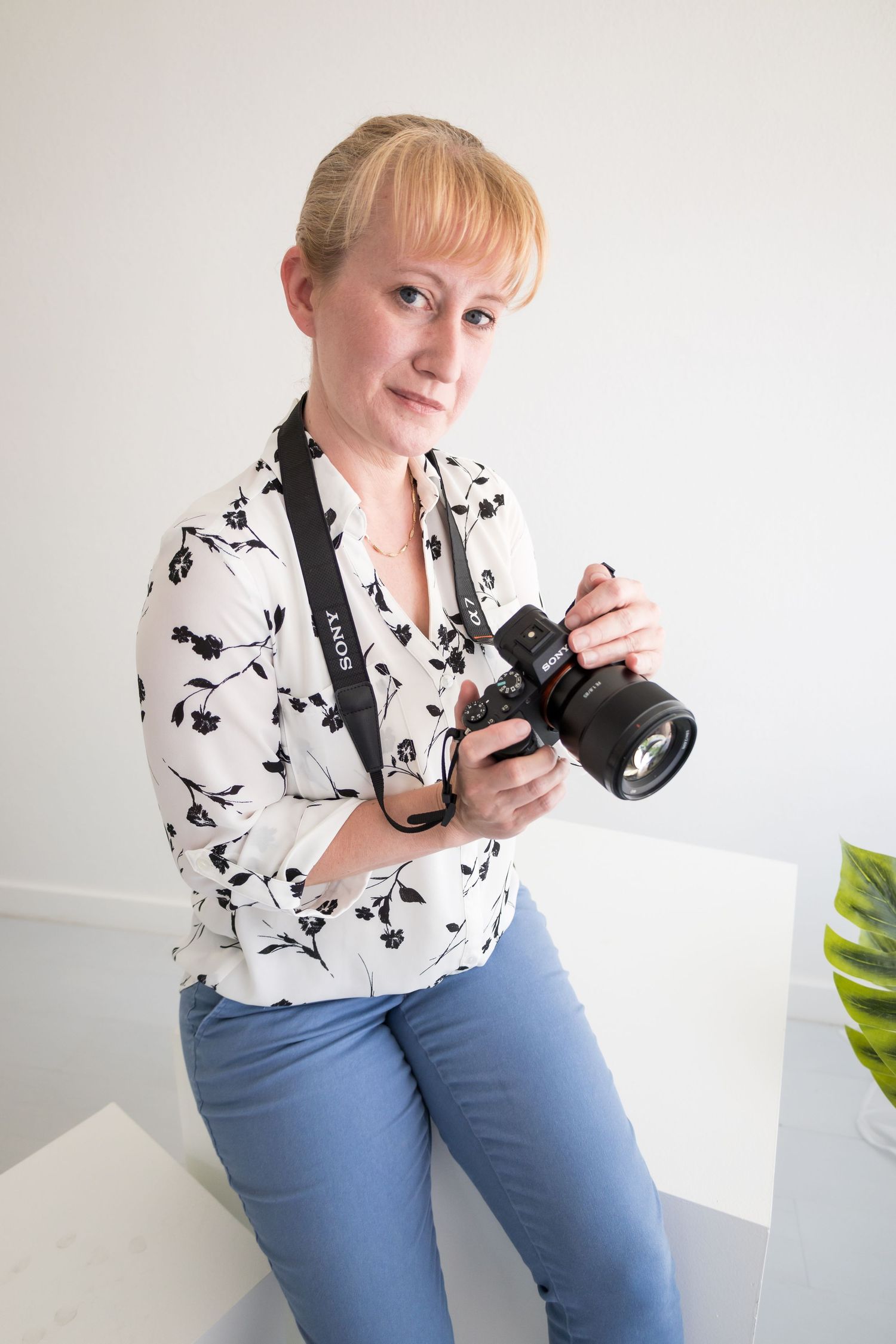 A photographer in a patterned blouse holds a professional camera against a white wall.
