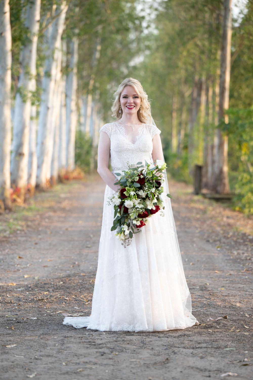Bride in lace wedding gown holds cascading burgundy and white bouquet in birch tree forest.