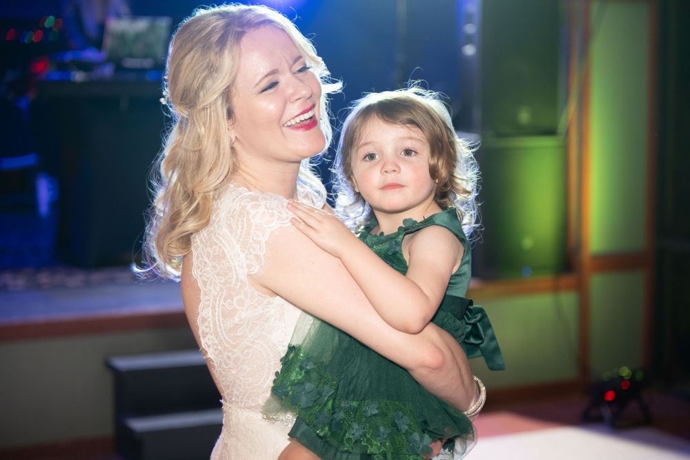 Dancing at wedding reception with bride in white lace gown holding young child in green dress.