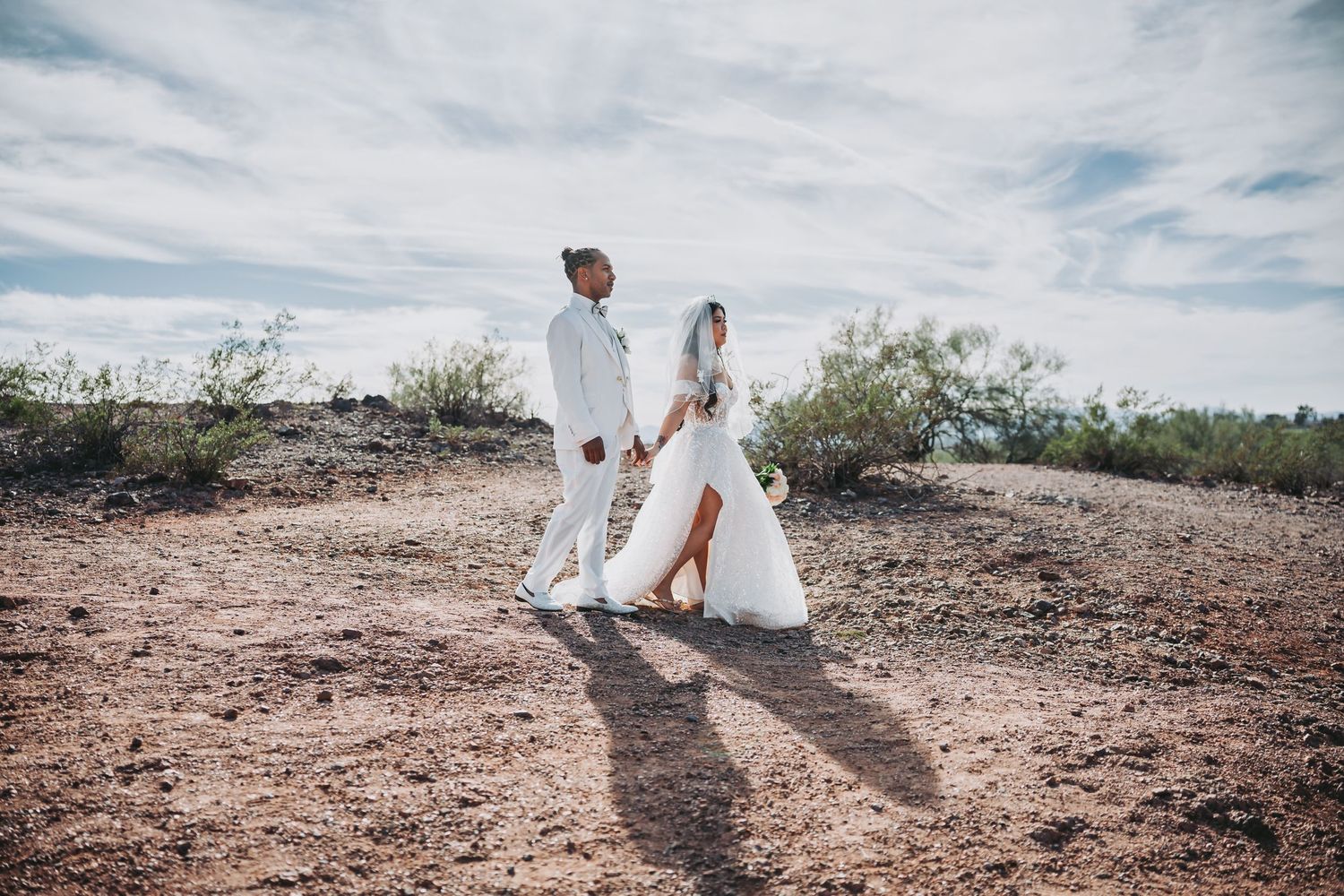 A couple in white attire walks through a desert landscape on their wedding day.