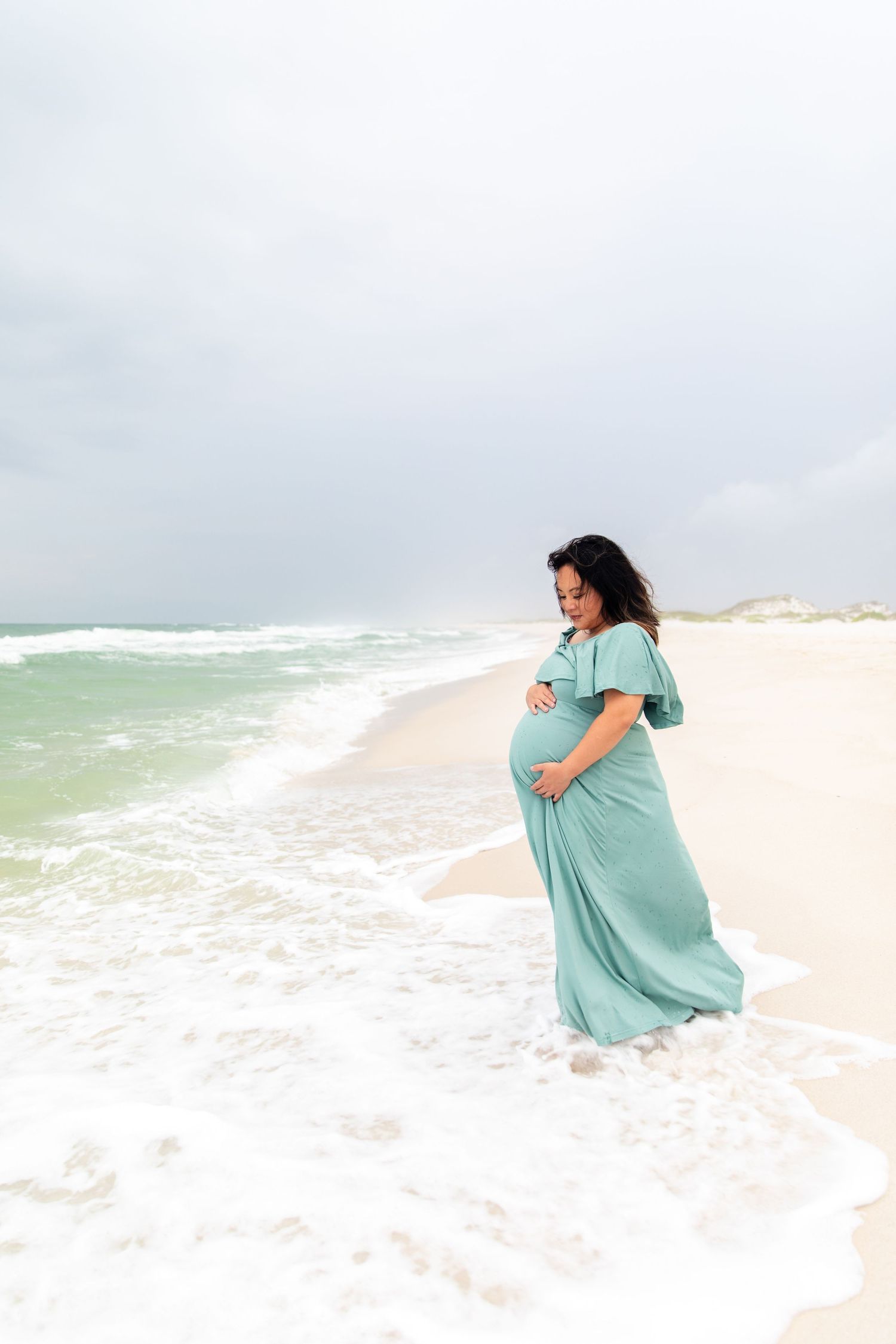 A pregnant woman in a teal dress stands in ocean waves on a cloudy beach day.