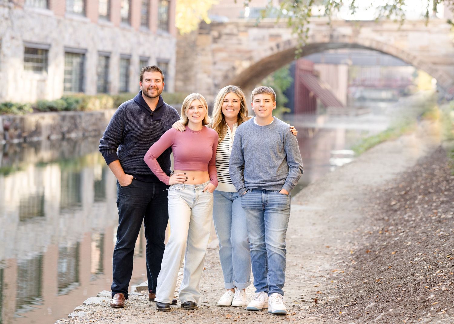 A group stands casually on a footpath alongside a scenic canal with a stone bridge in the background.