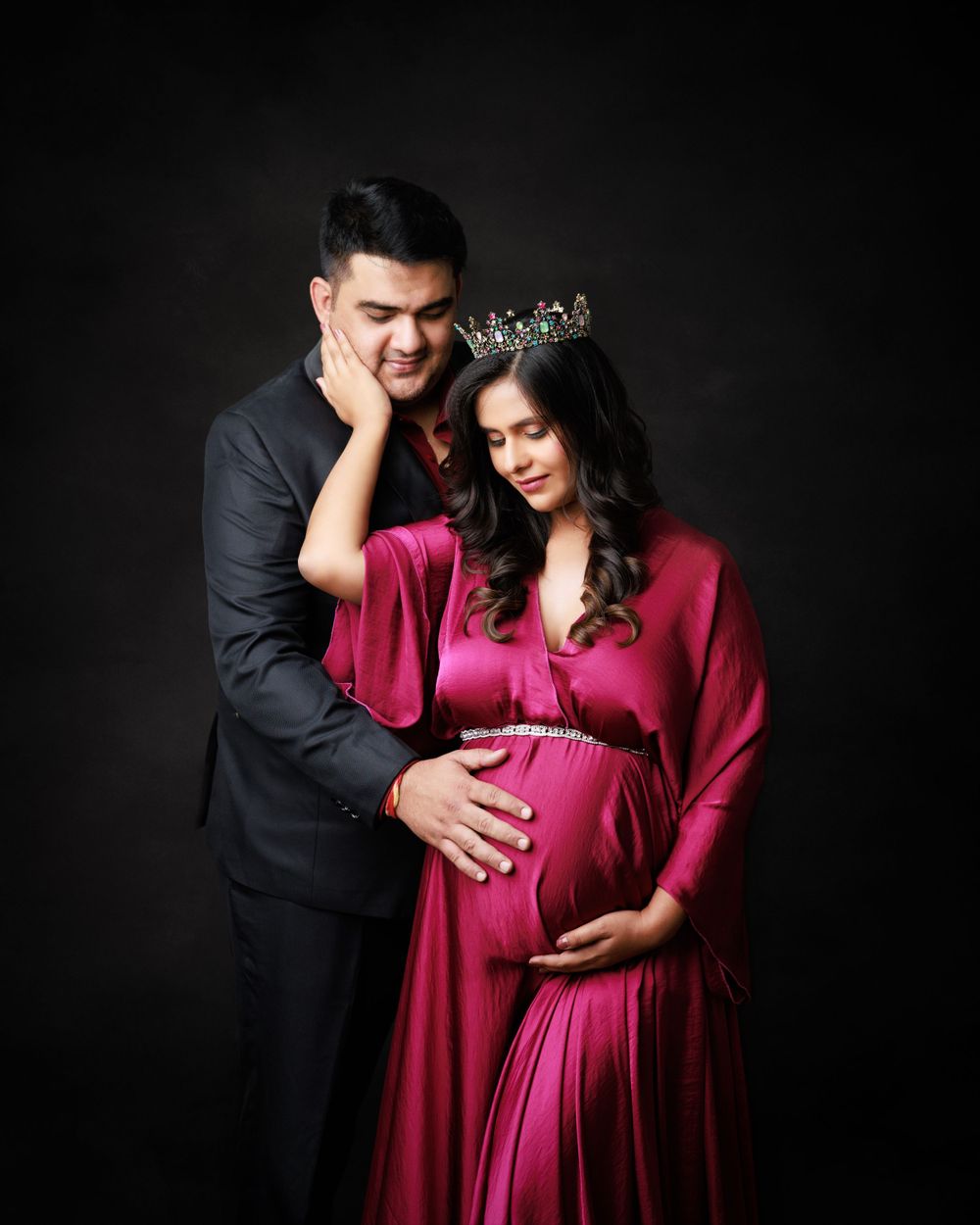 Intimate maternity portrait of couple in formal attire against dark background.