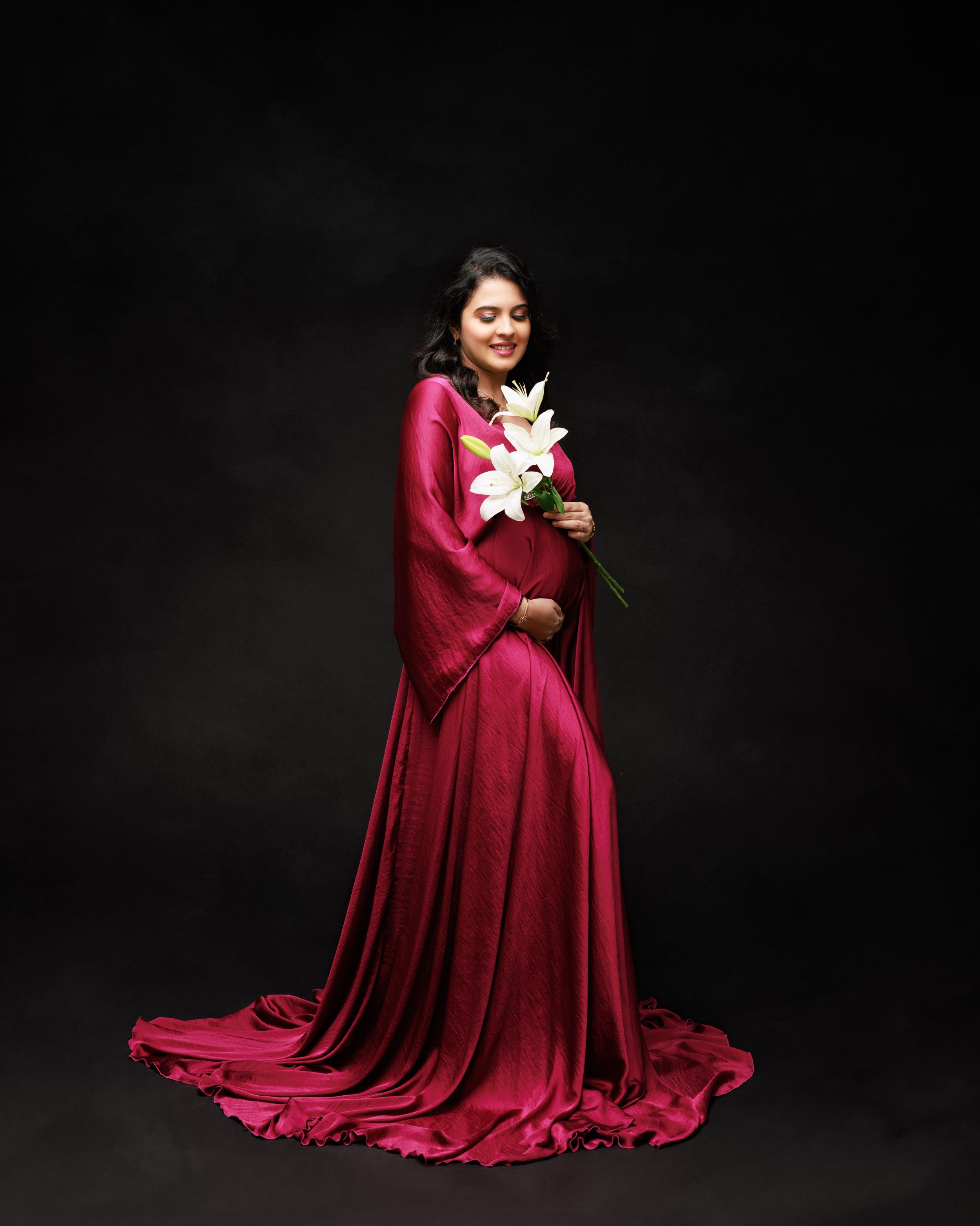 Elegant maternity photo featuring a woman in flowing red gown holding white lilies against dark background.