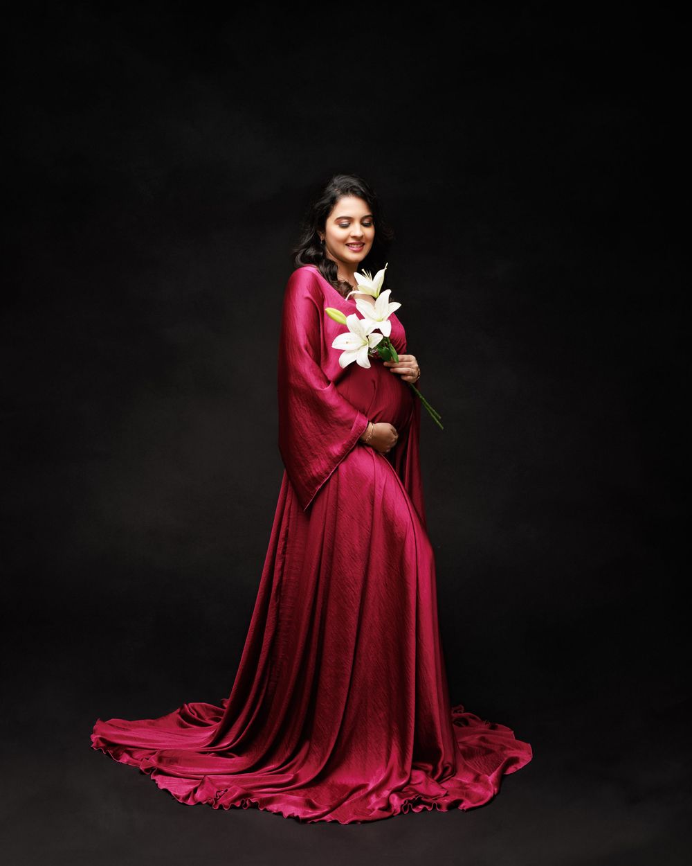 Elegant maternity photo featuring a woman in flowing red gown holding white lilies against dark background.