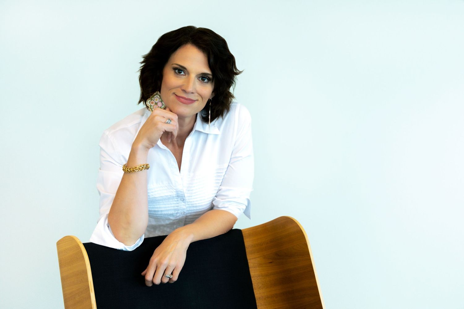 Professional headshot of woman in white shirt seated on wooden chair against light background.