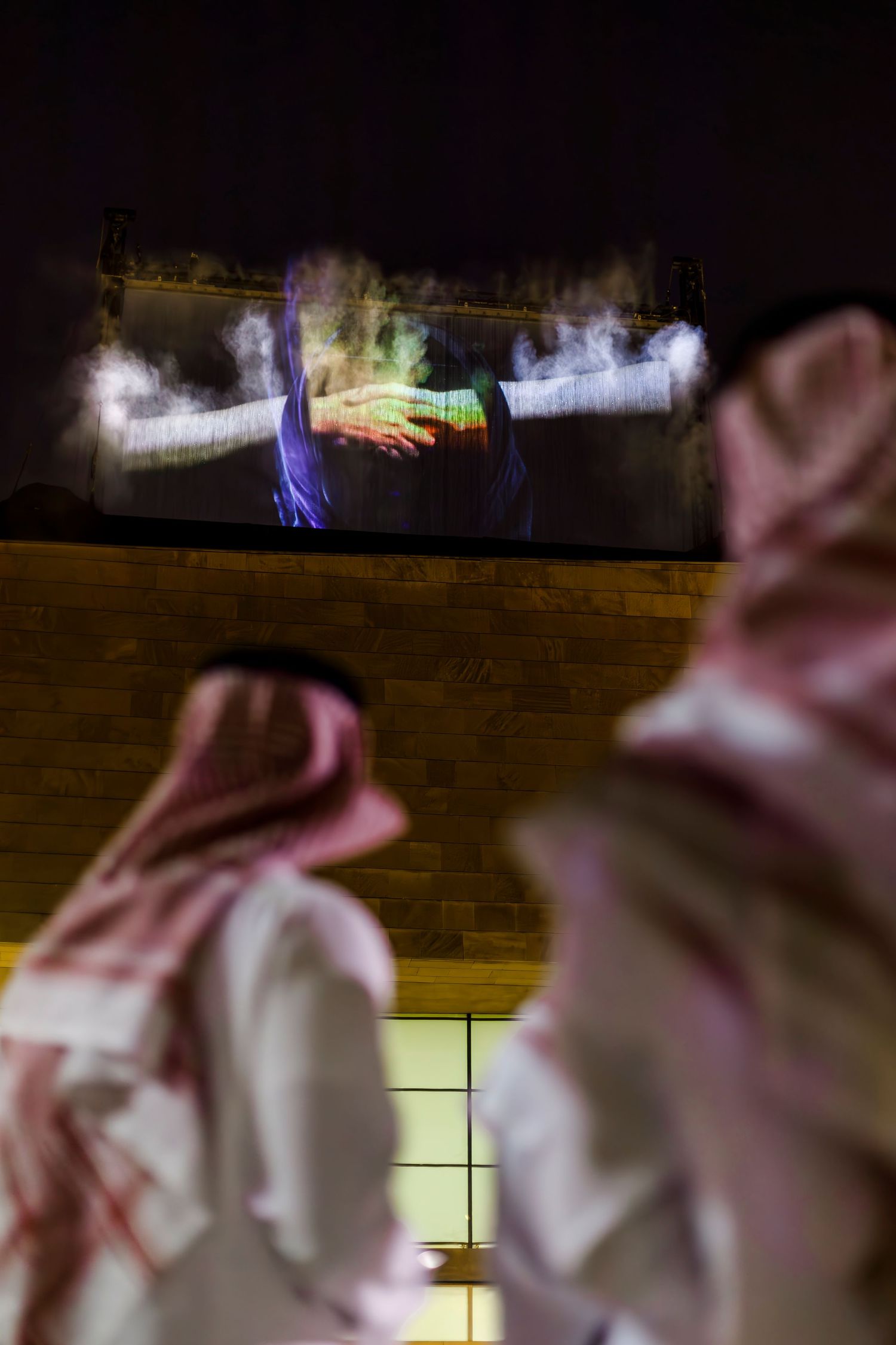 Colorful light projections illuminate a wall as spectators watch in traditional Middle Eastern dress.
