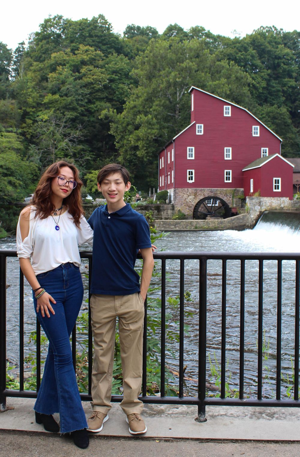 Two young people pose by metal railing with a red mill and waterfall in background.