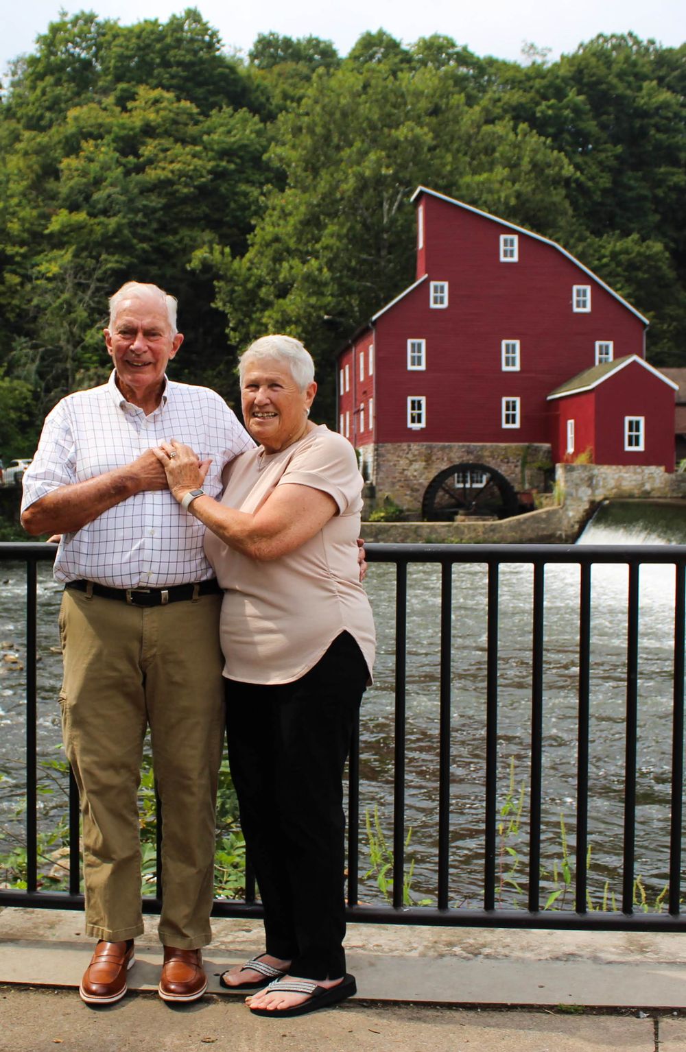 Couple stands together near railing with historic red mill and waterfall in the background.