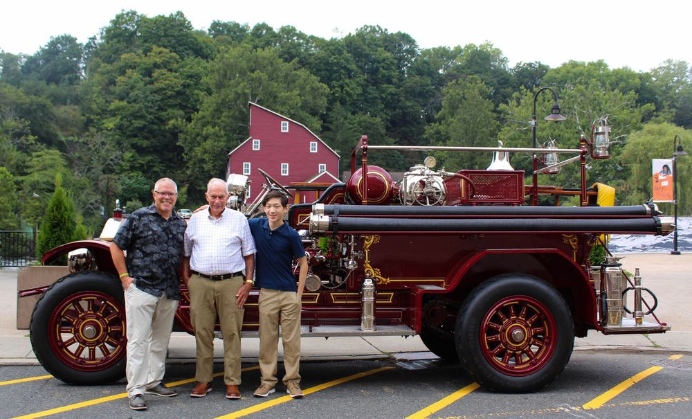 Three people stand beside a restored antique fire engine with a red barn visible in the background.