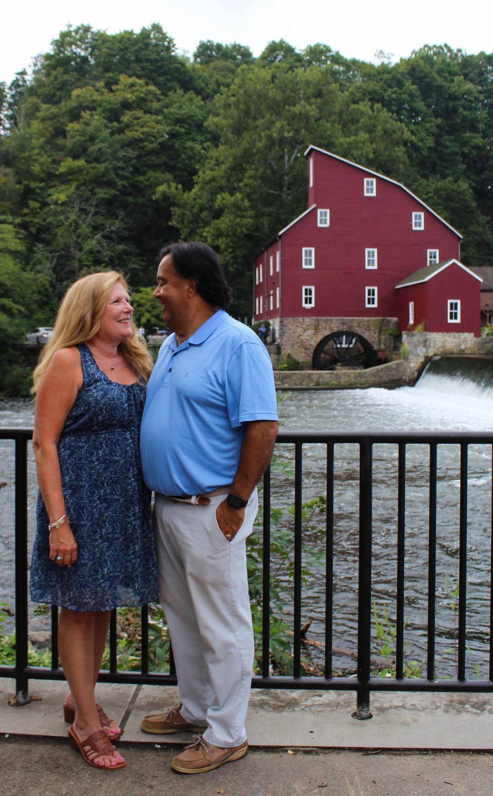 A couple poses in front of a historic red mill building with rushing water and green trees in the background.