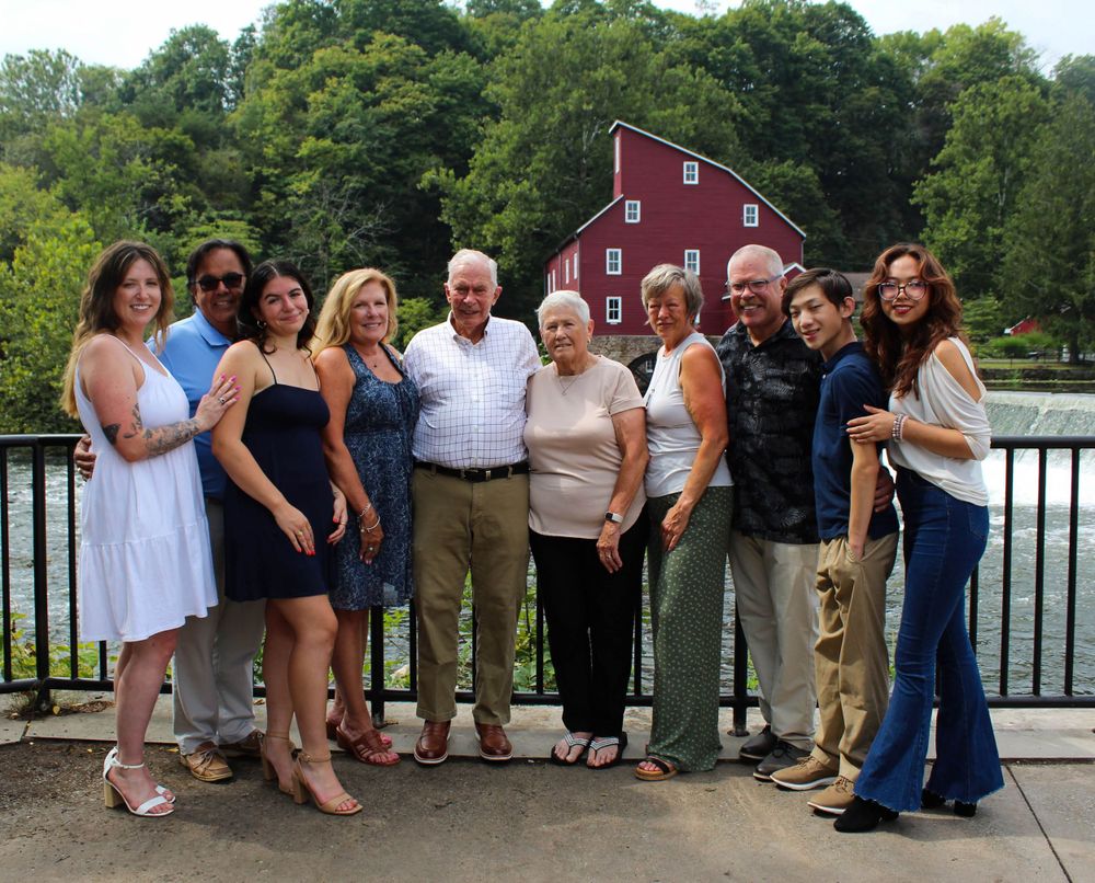 Extended family gathers for group photo by railing with red mill and water behind them.