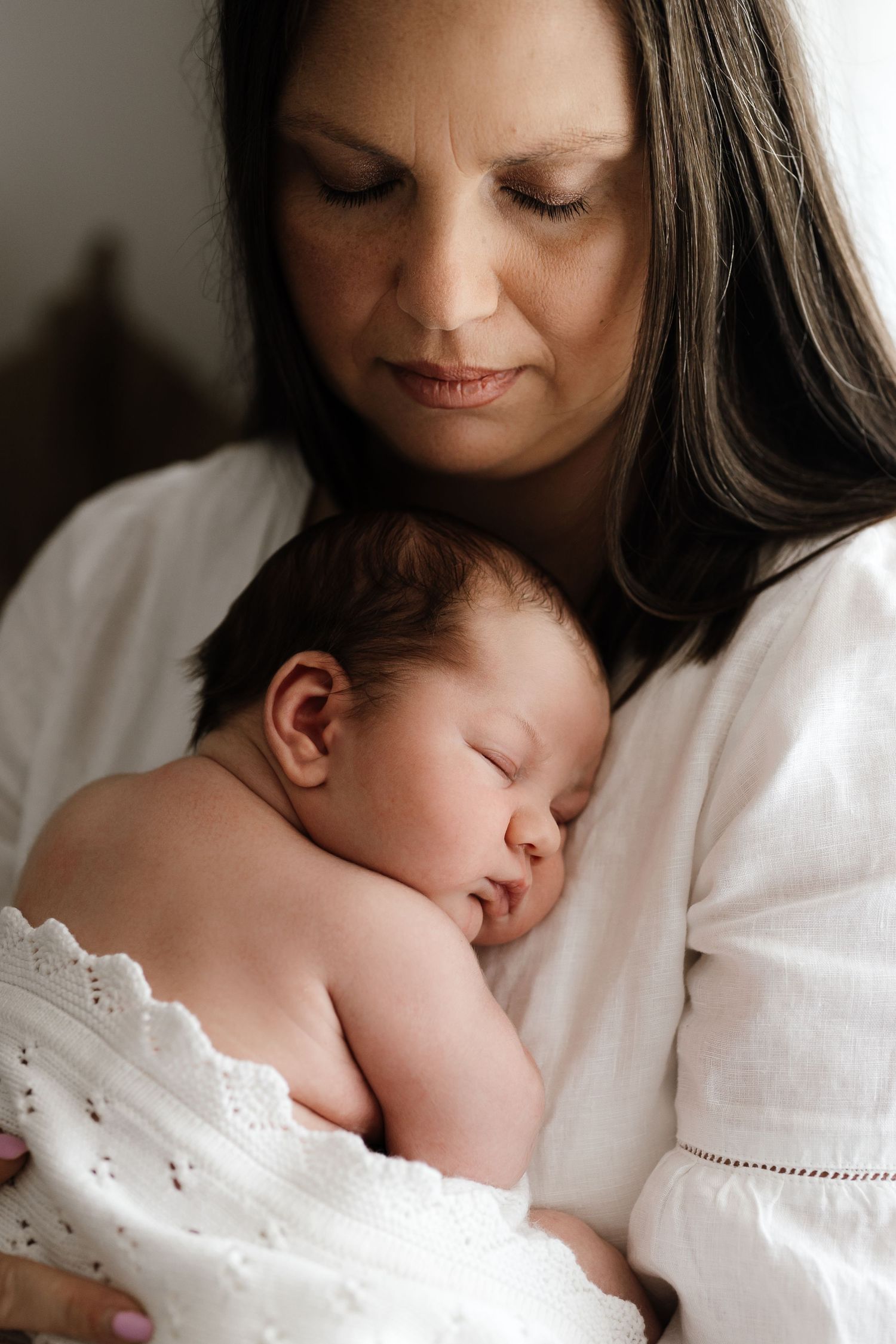 Tender moment of a dark-haired mother in white dress cradling her sleeping newborn baby.