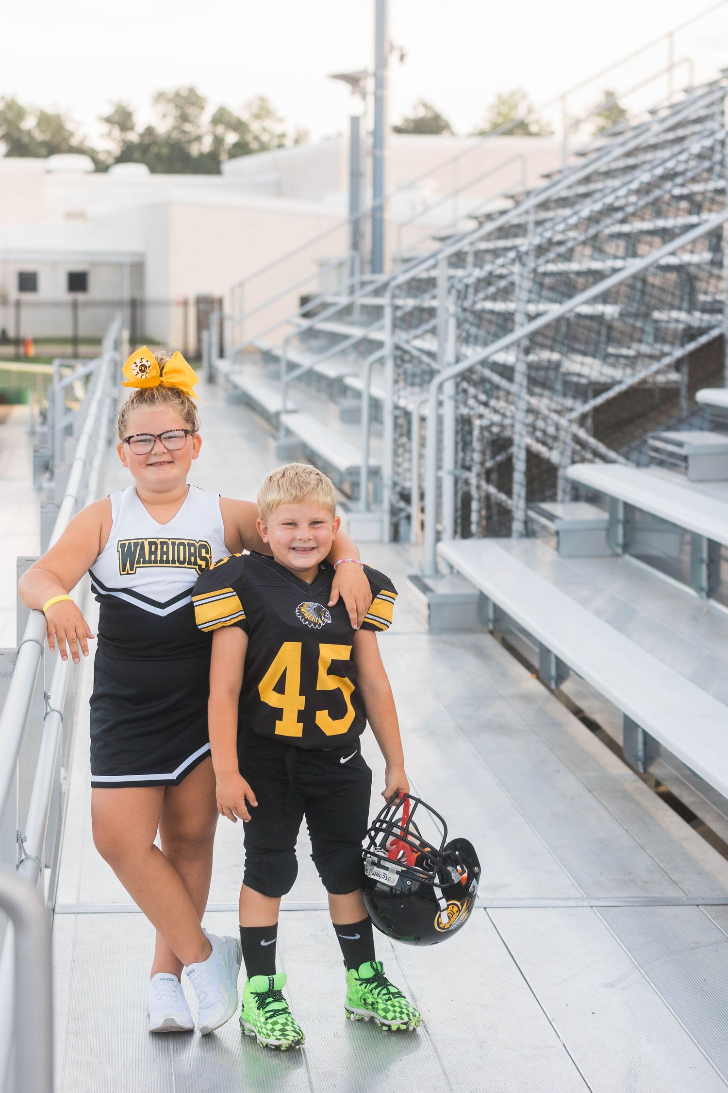 Two youth athletes in football and cheerleading uniforms pose together on stadium bleachers.