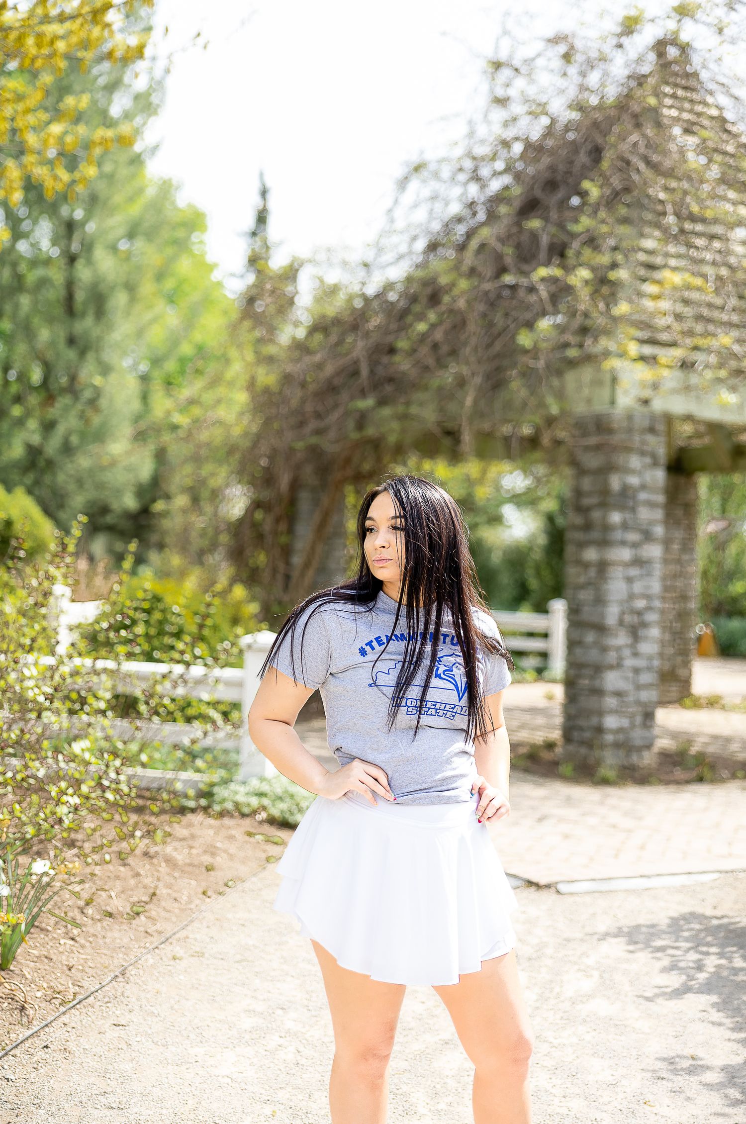 A woman in a casual gray t-shirt and white skirt poses near a stone archway in a sunny garden setting.
