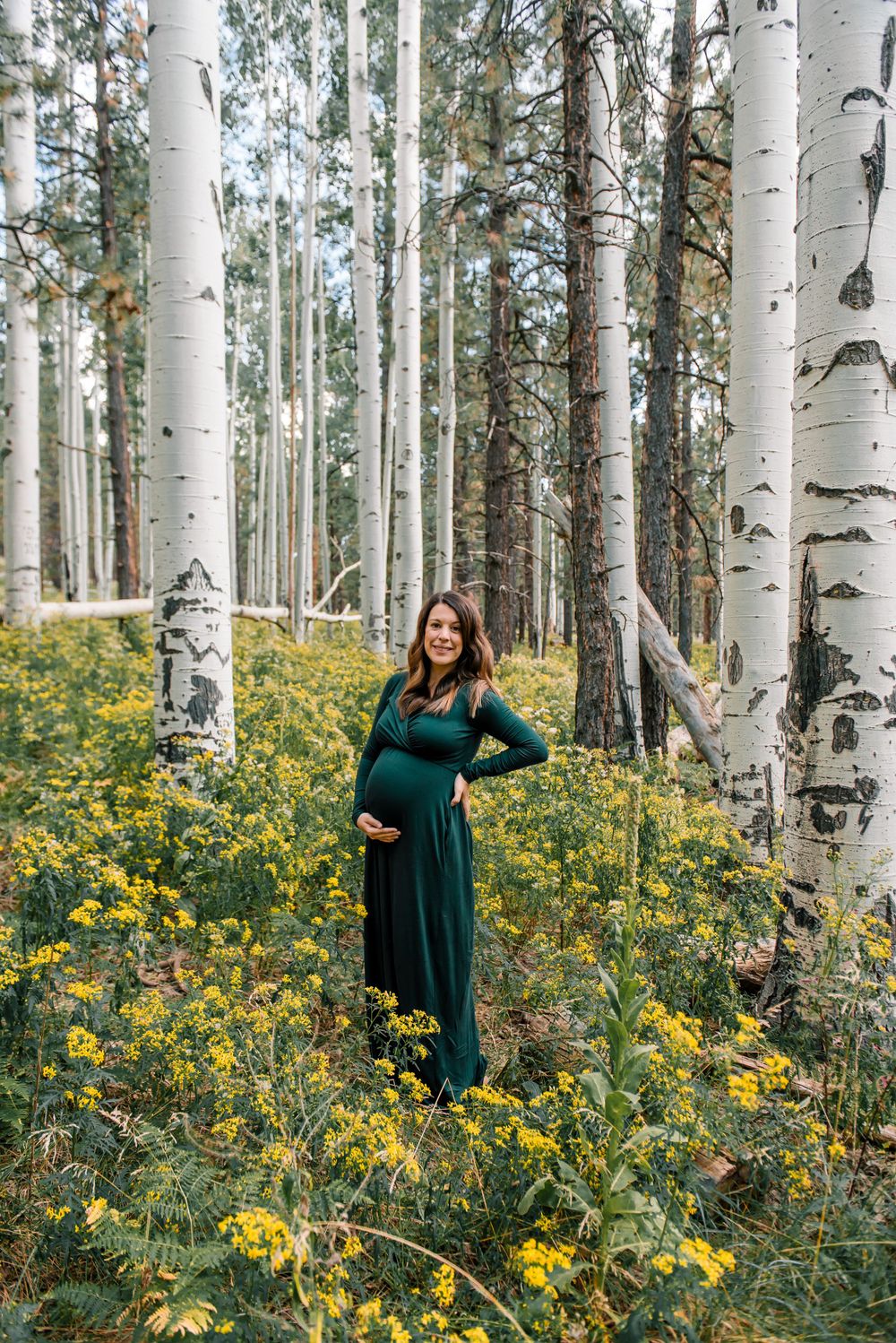 A couple poses in a forest of white birch trees with yellow wildflowers blooming beneath.