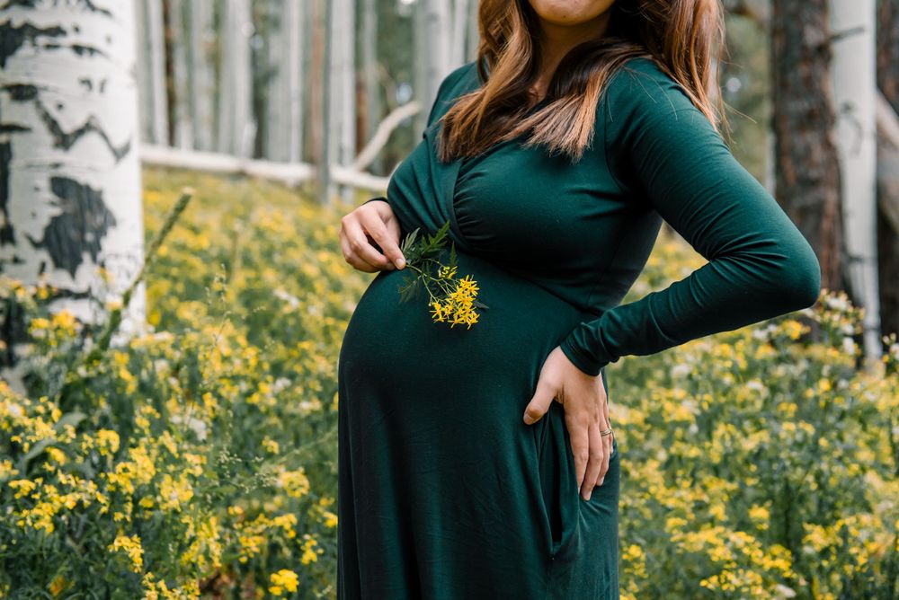 A woman in an emerald green dress cradles her baby bump in a field of yellow wildflowers.
