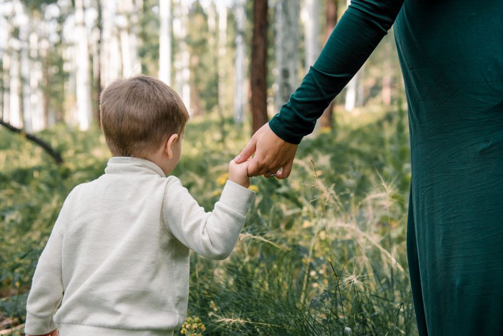 A small child holds hands with someone wearing a green dress in a forest setting.