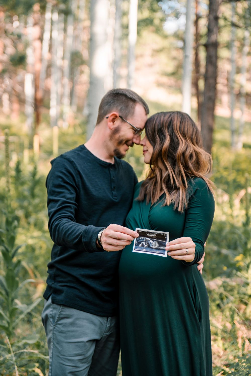 A couple in dark clothing looks at a sonogram photo together in a wooded setting.