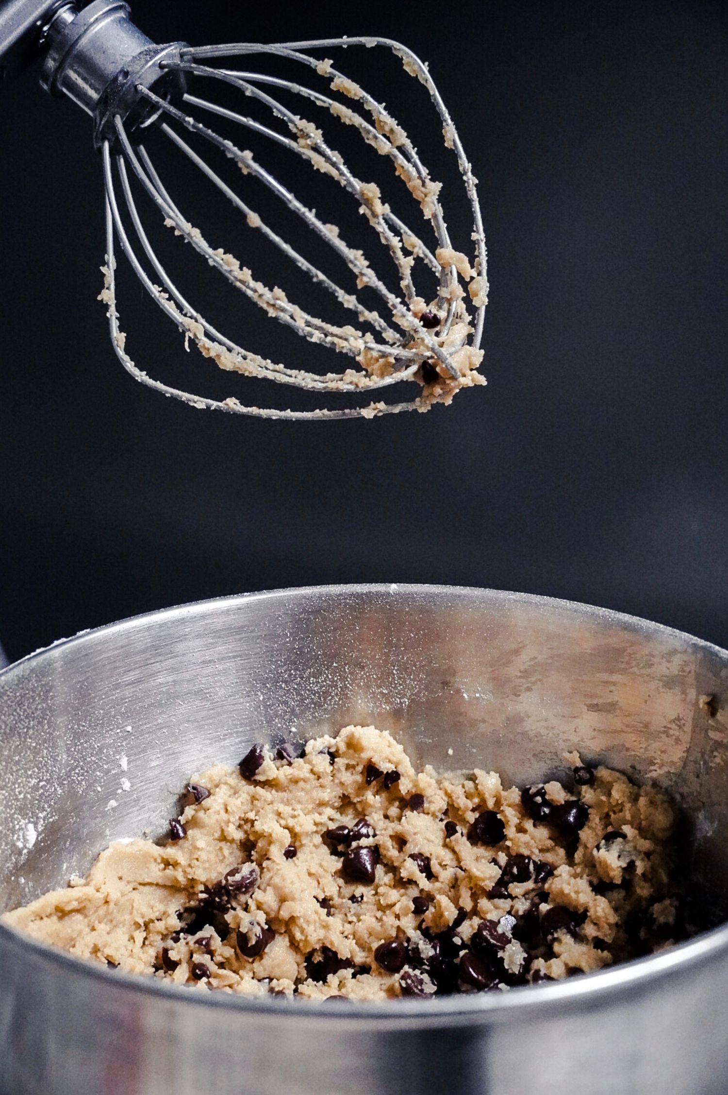 Whisk attachment covered in cookie dough mixture hovers over mixing bowl with chocolate chip cookie batter.