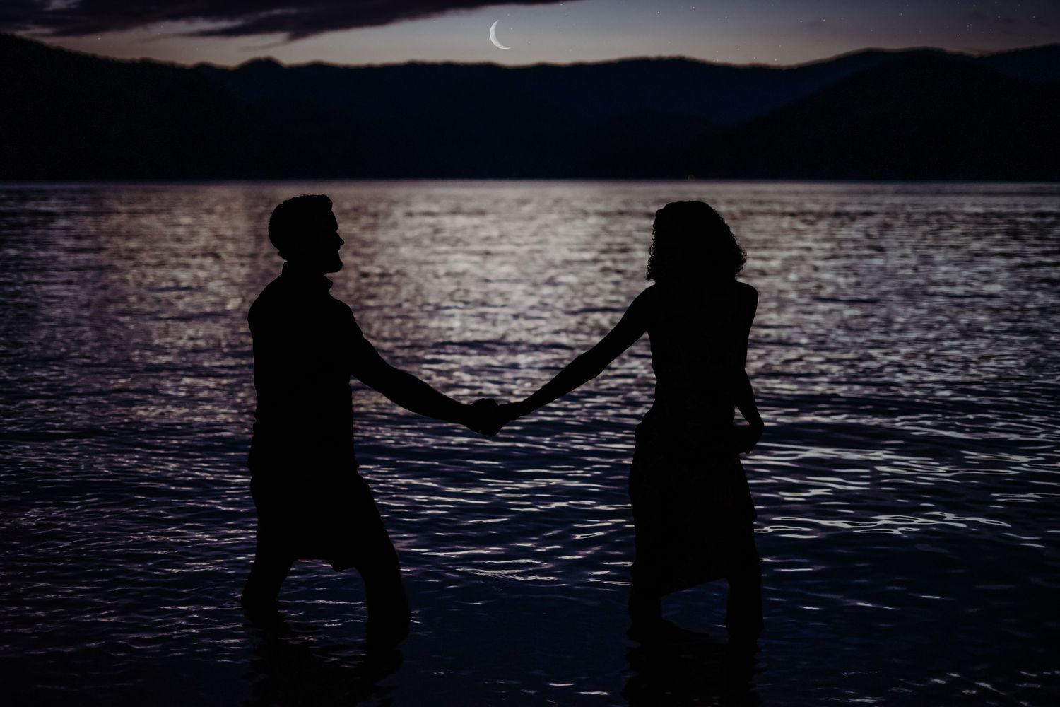 Silhouettes of figures by moonlit lake creating dramatic nighttime scene.