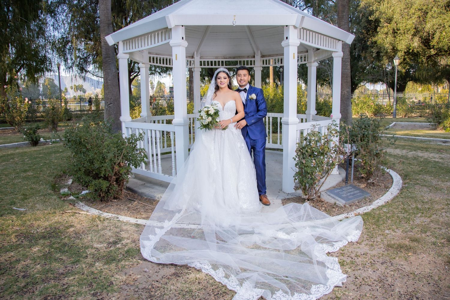 Bride and groom posing together in front of a white gazebo in a lush garden on their wedding day.