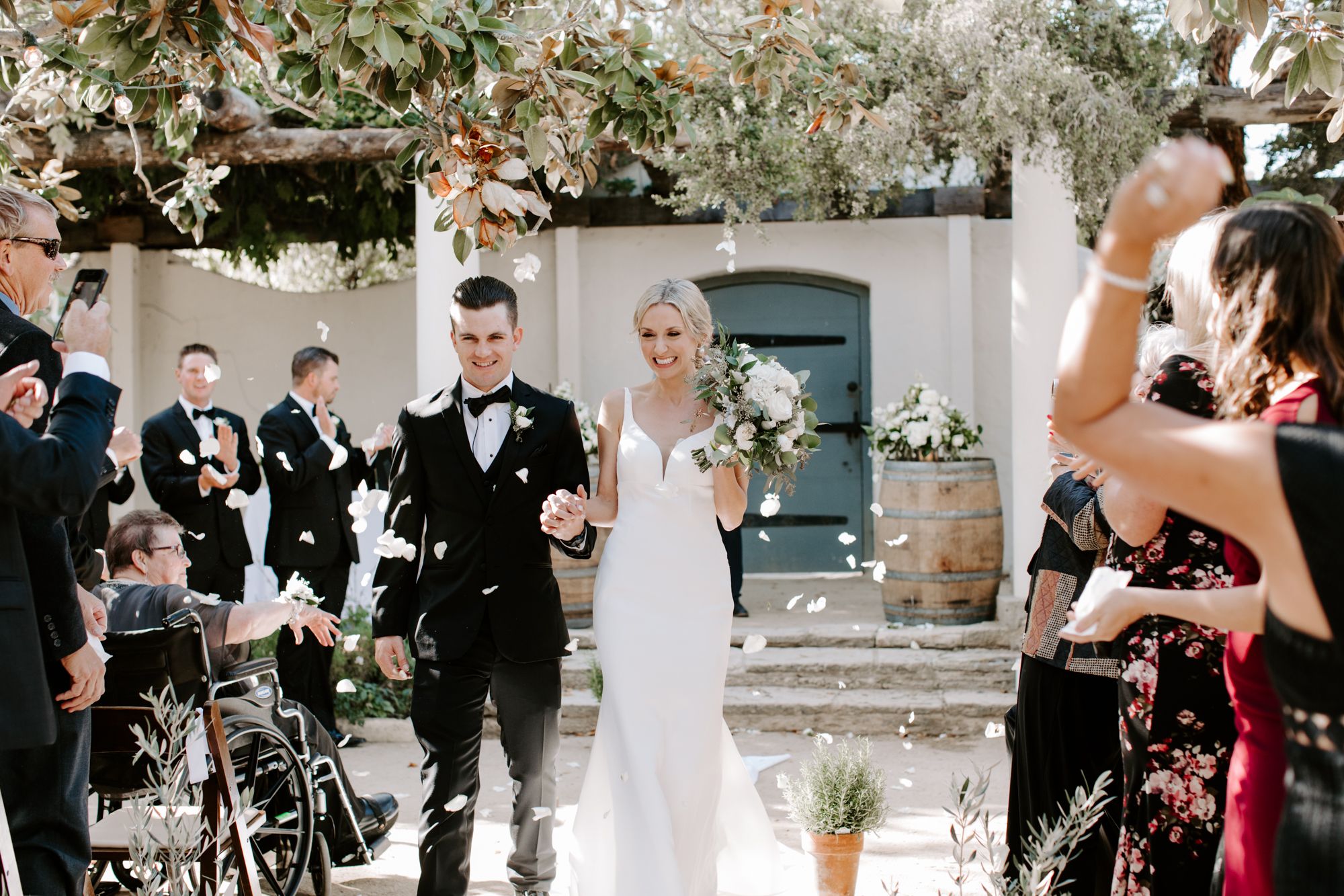 Wedding guests throw flower petals as newlyweds exit their ceremony under floral arrangements.
