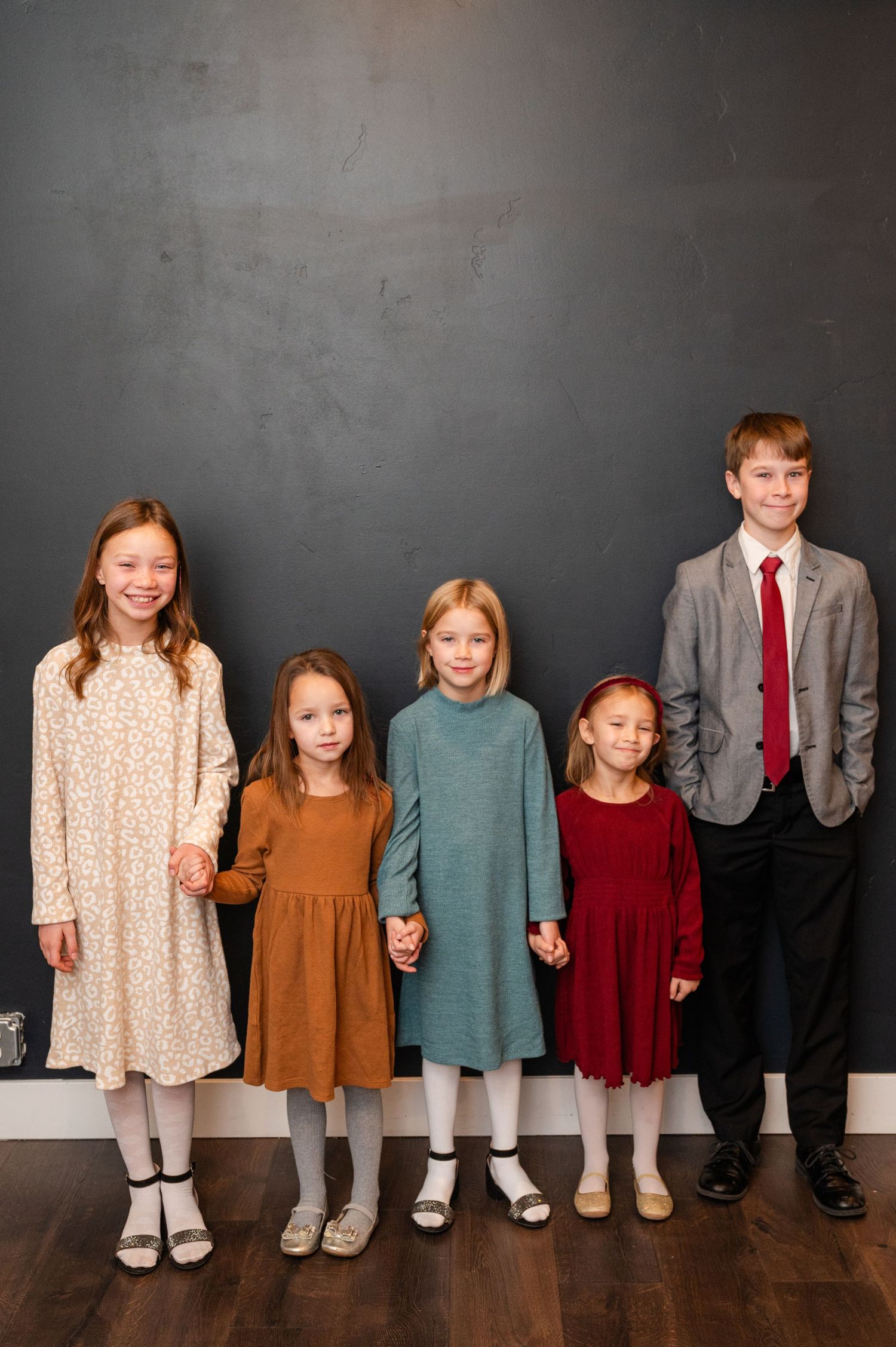 Five children standing in line against dark wall wearing coordinated dresses and suits.