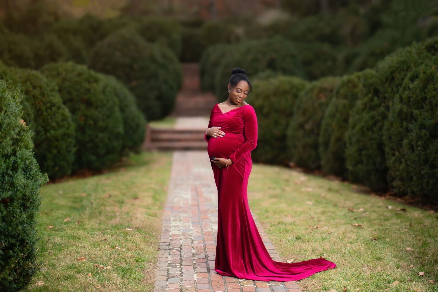Person wearing red formal gown poses in garden with manicured hedges during maternity shoot.