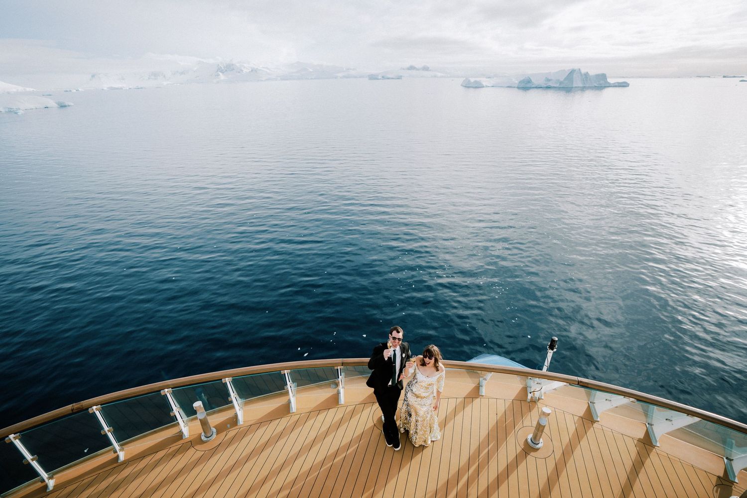 A couple standing on the bow deck of a ship with icy waters and snow-capped mountains in the background.