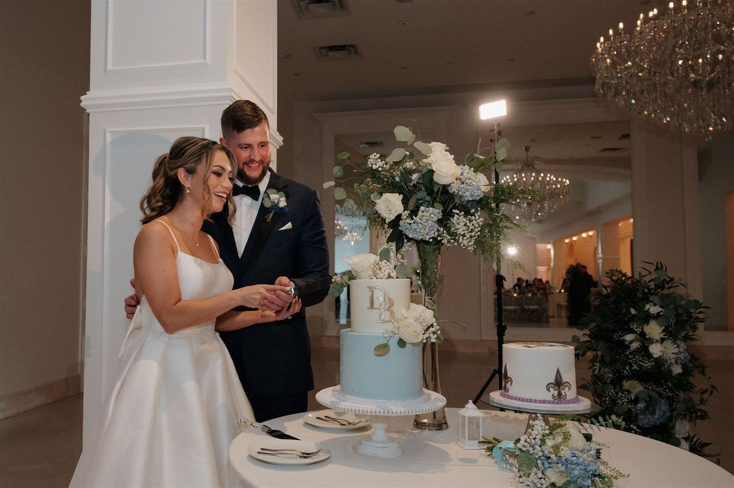 A couple shares a happy moment while cutting their elegant white wedding cake decorated with greenery at their reception.