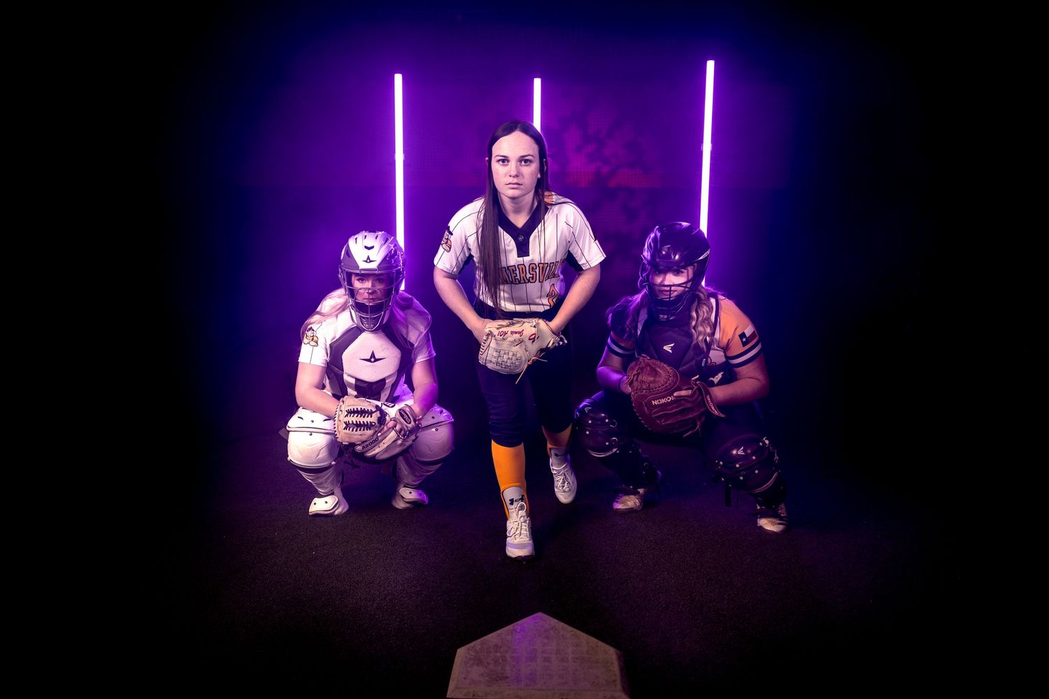 Three softball players pose in dramatic lighting with neon purple effects.