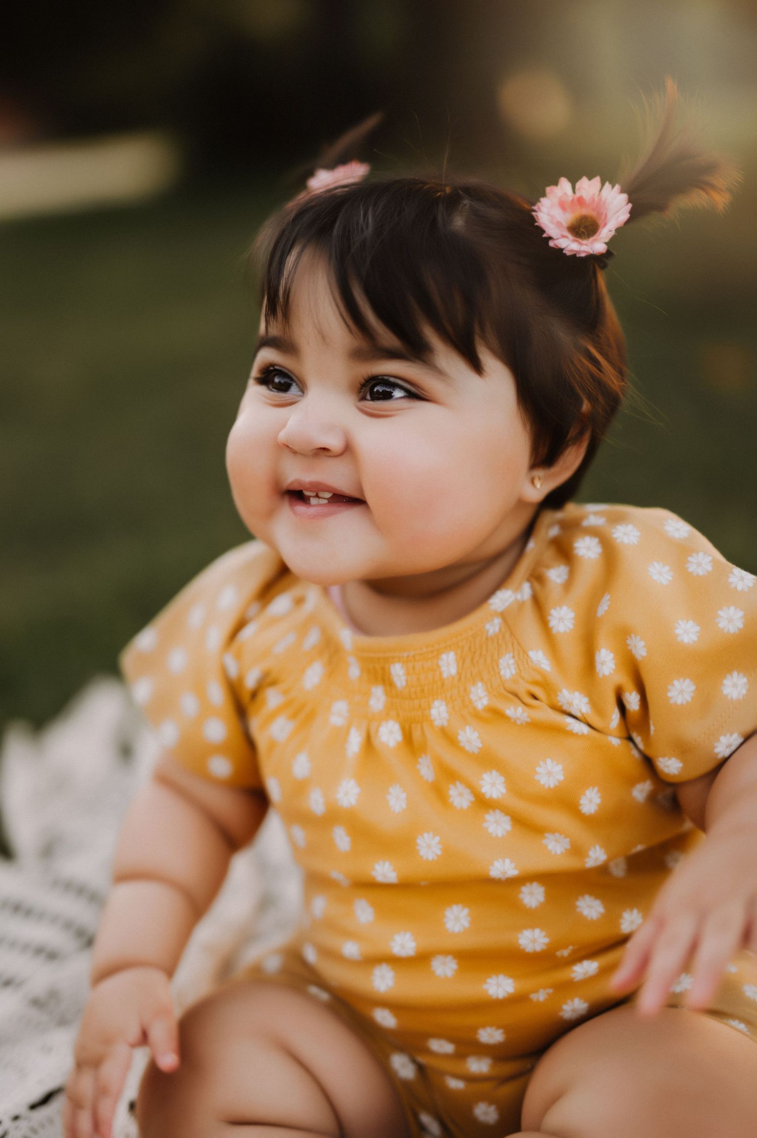 Baby in yellow polka dot outfit with flower hair clip smiling brightly outdoors.