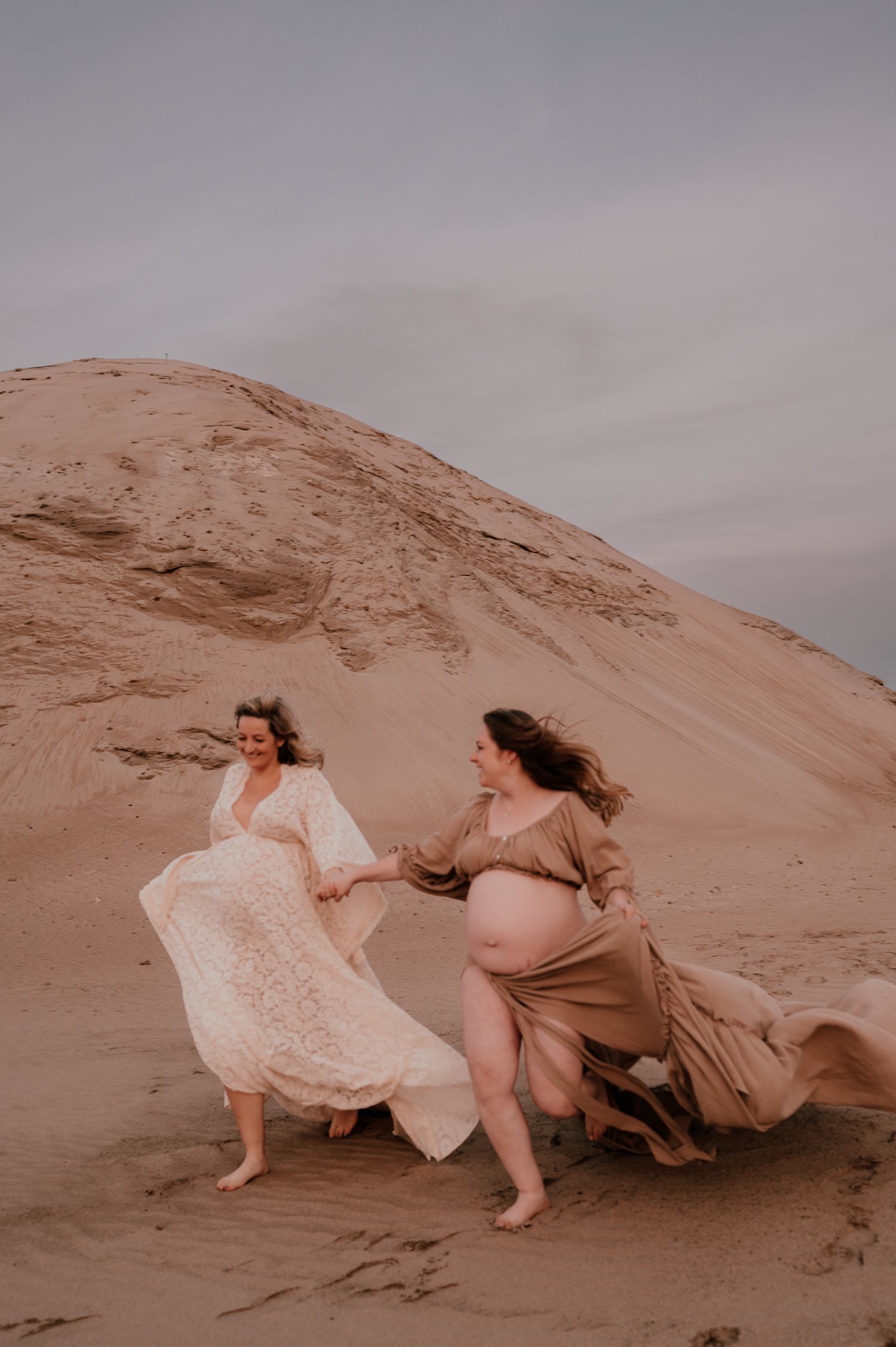 Two people in flowing dresses running across sand dunes at sunset.
