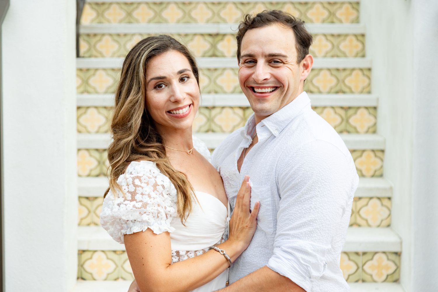 Engagement photo of couple in white attire against a decorative tiled background.