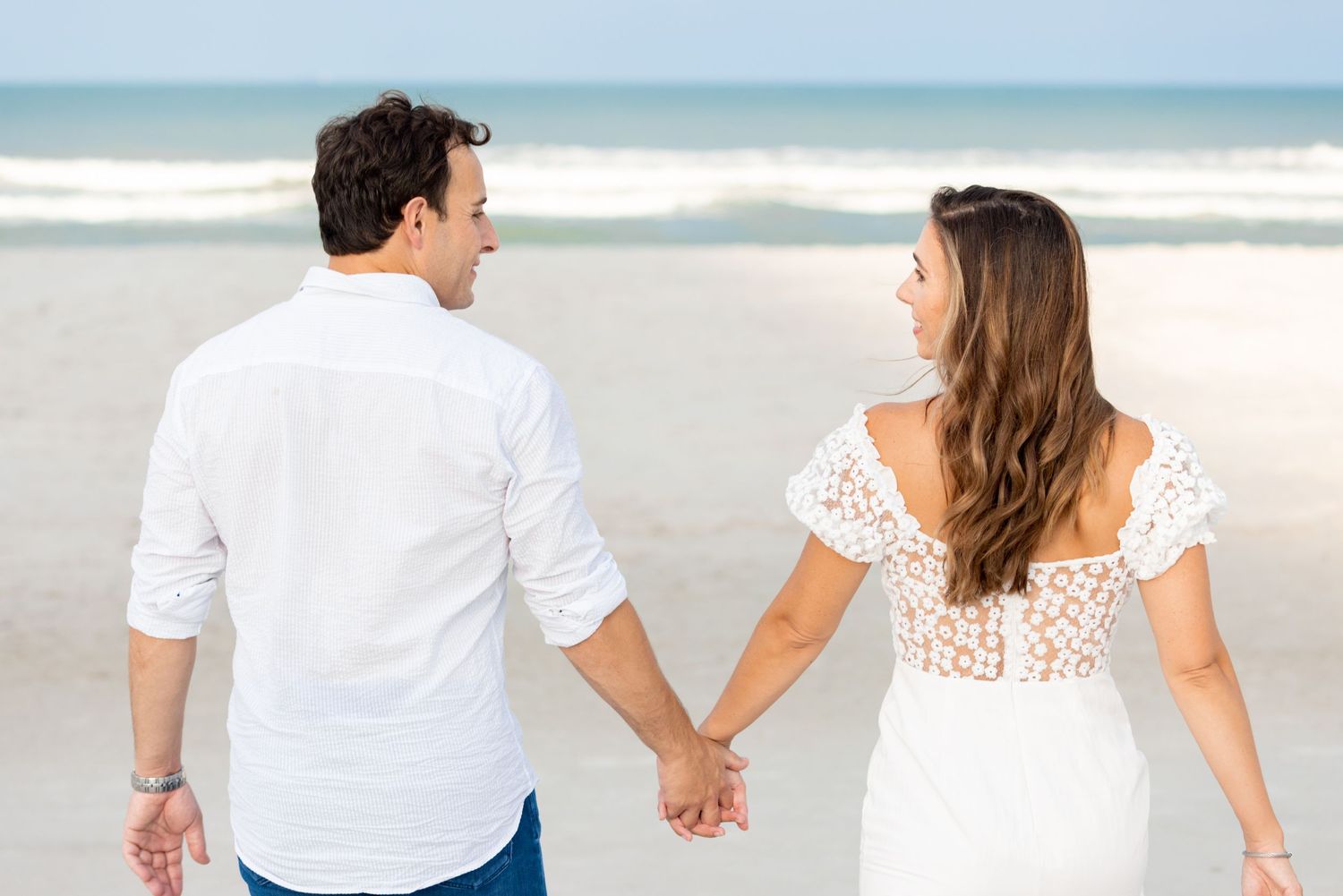 Couple walking hand in hand on a beach wearing coordinated white outfits.