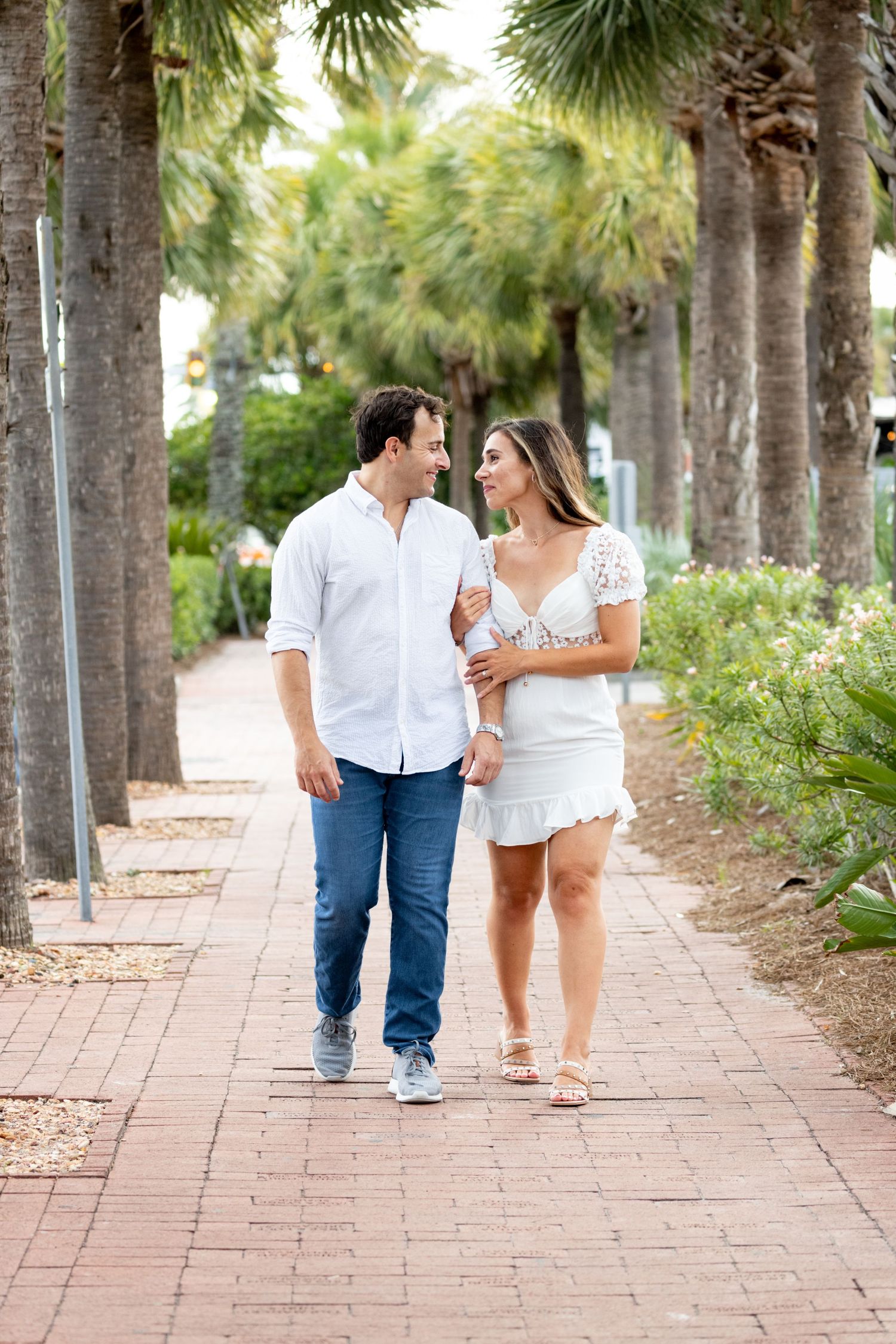 Couple strolls along palm-lined brick pathway in coordinated white outfits.
