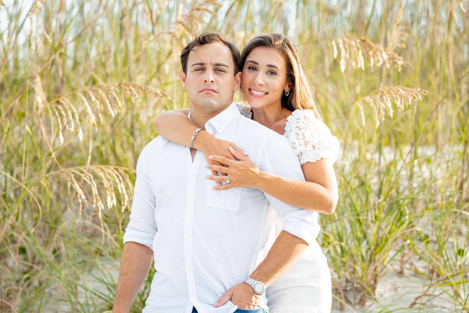 Couple in white outfits embrace in tall beach grass with soft natural lighting.