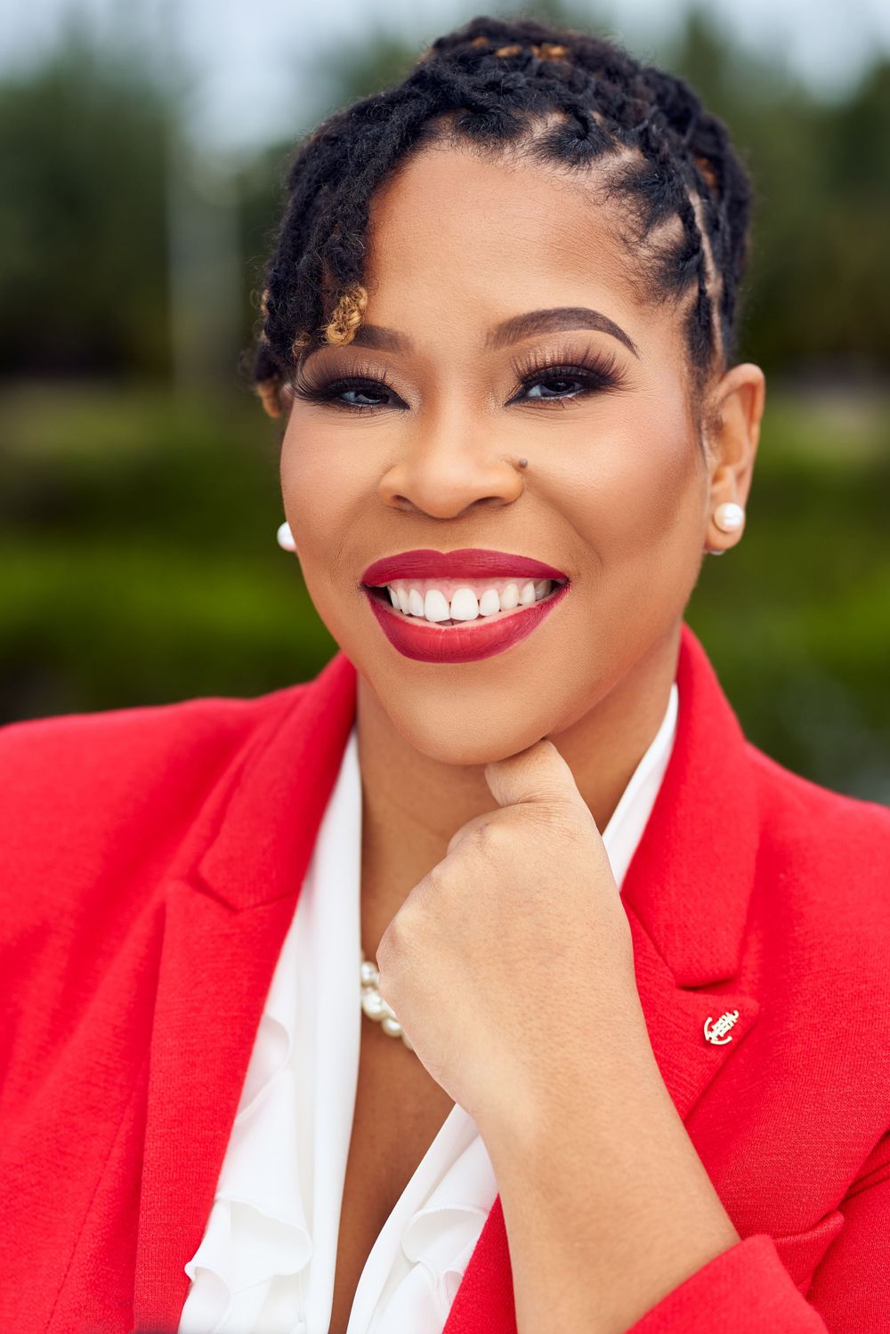Professional headshot of a woman in a red blazer and white top with braided hair and bright smile against blurred background.