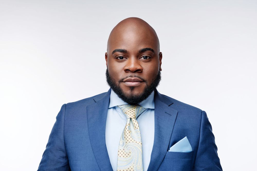 Professional headshot of man in blue suit with patterned tie and pocket square against light background.