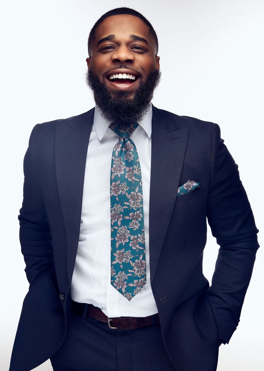 Professional headshot of man with full beard wearing navy suit and floral tie against white background.