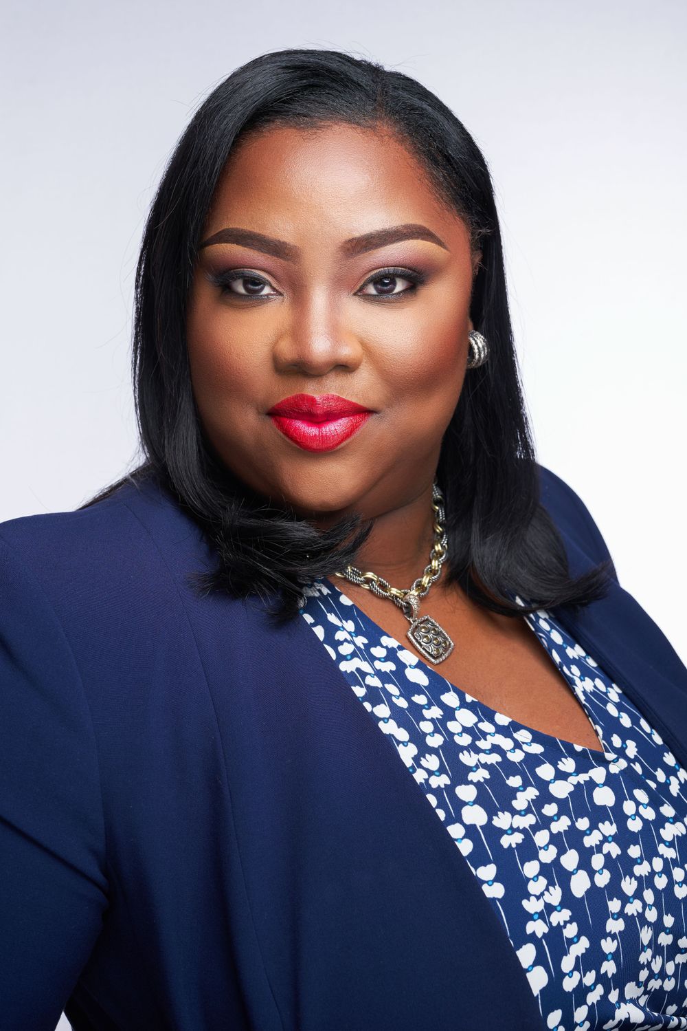Professional portrait of woman in navy blazer and floral dress with statement necklace against light background.