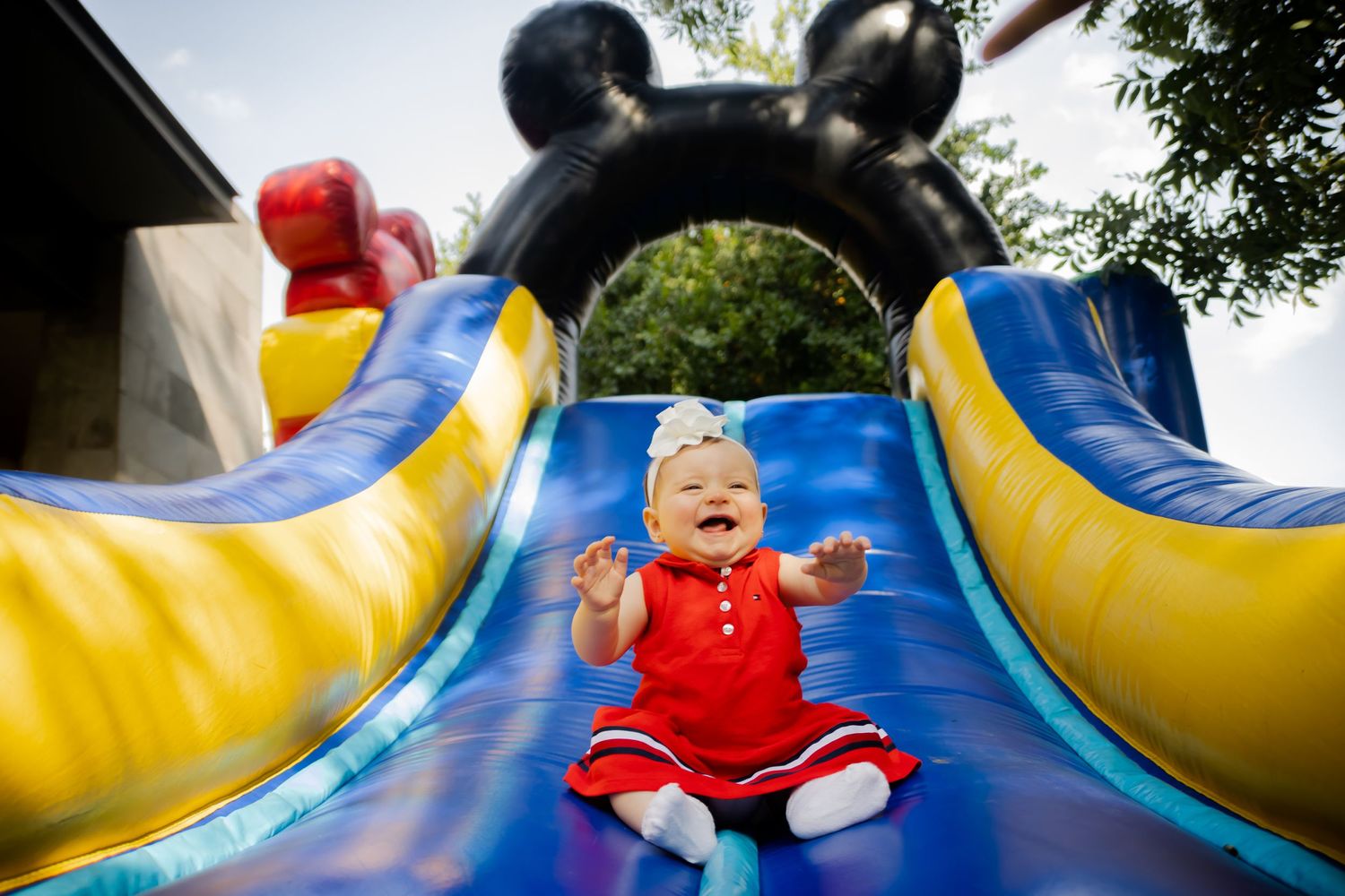 Happy baby in red dress slides down a colorful inflatable bounce house slide outdoors.