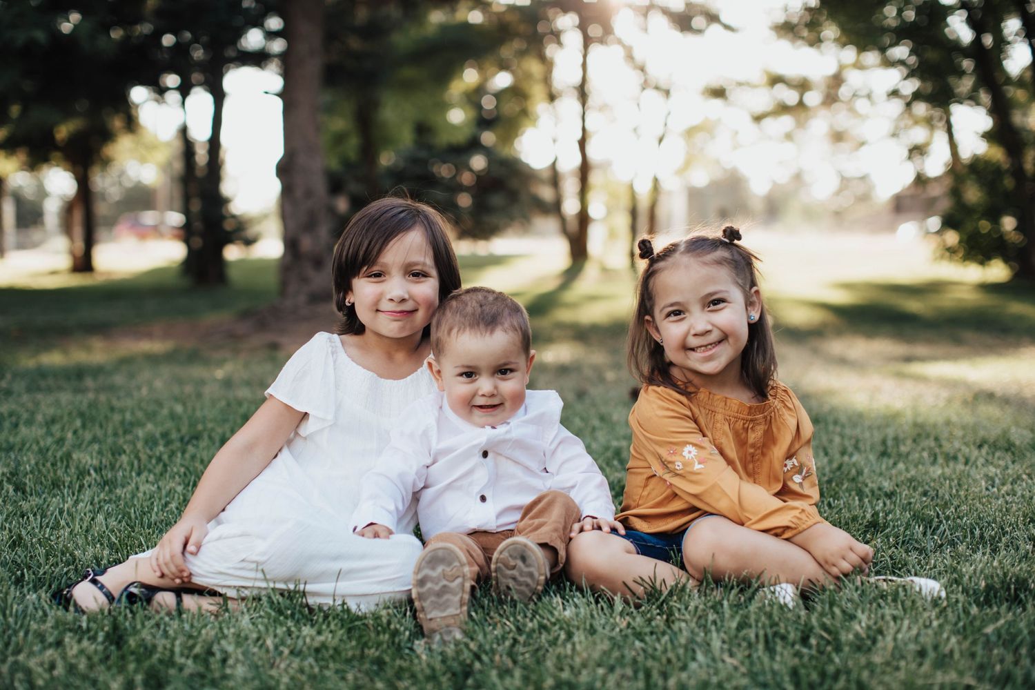 Tucson children’s portrait of three young sisters sitting in the grass smiling in natural, warm light.